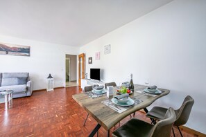 A dining area with a wooden table set for four, a grey sofa, and a TV in the background. - Lido Apartment Locarno With Balcony and Parking (Locarno)