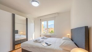 A modern hotel room with a large bed, a mirror, a window with a view of buildings, and a bedside lamp. - Lido Apartment Locarno With Balcony and Parking (Locarno)