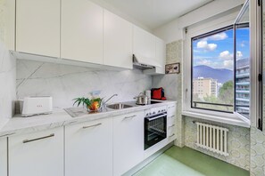 Una cocina con armarios blancos, horno empotrado, ventana con vistas a edificios y montañas, y un radiador.