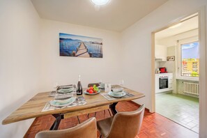 A dining area with a wooden table set for two, a painting on the wall, and a view of the kitchen through an open door. - Lido Apartment Locarno With Balcony and Parking (Locarno)