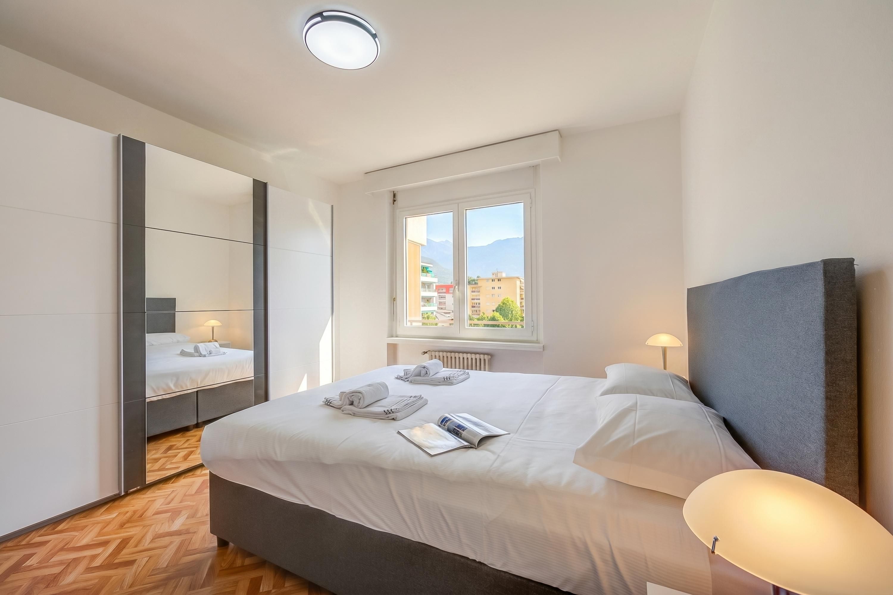 A modern hotel room with a large bed, a mirror, a window with a view of buildings, and a bedside lamp.