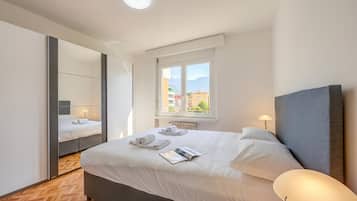 A modern hotel room with a large bed, a mirror, a window with a view of buildings, and a bedside lamp.
