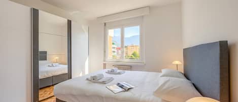 Una habitación de hotel moderna con una cama grande, un espejo, una ventana con vistas a edificios y una lámpara de noche.