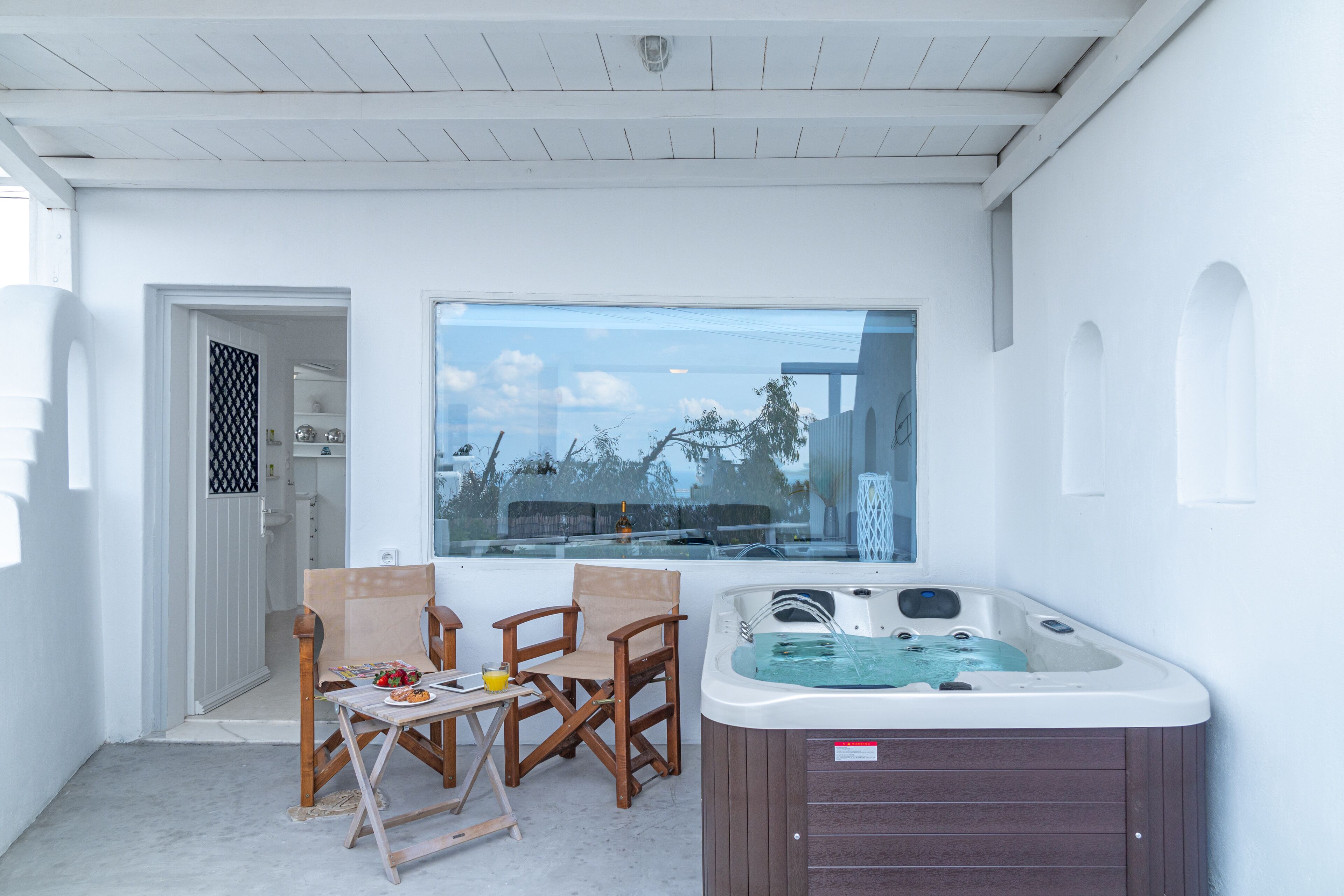 A room with a hot tub, wooden chairs, a table with a drink, and a view of the outdoors.