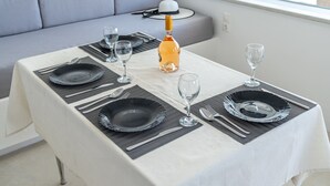 A dining table set with black plates, silverware, and a bottle of wine, with a gray sofa and a window in the background. - Bella View Mykonos Town Suites (Mykonos)