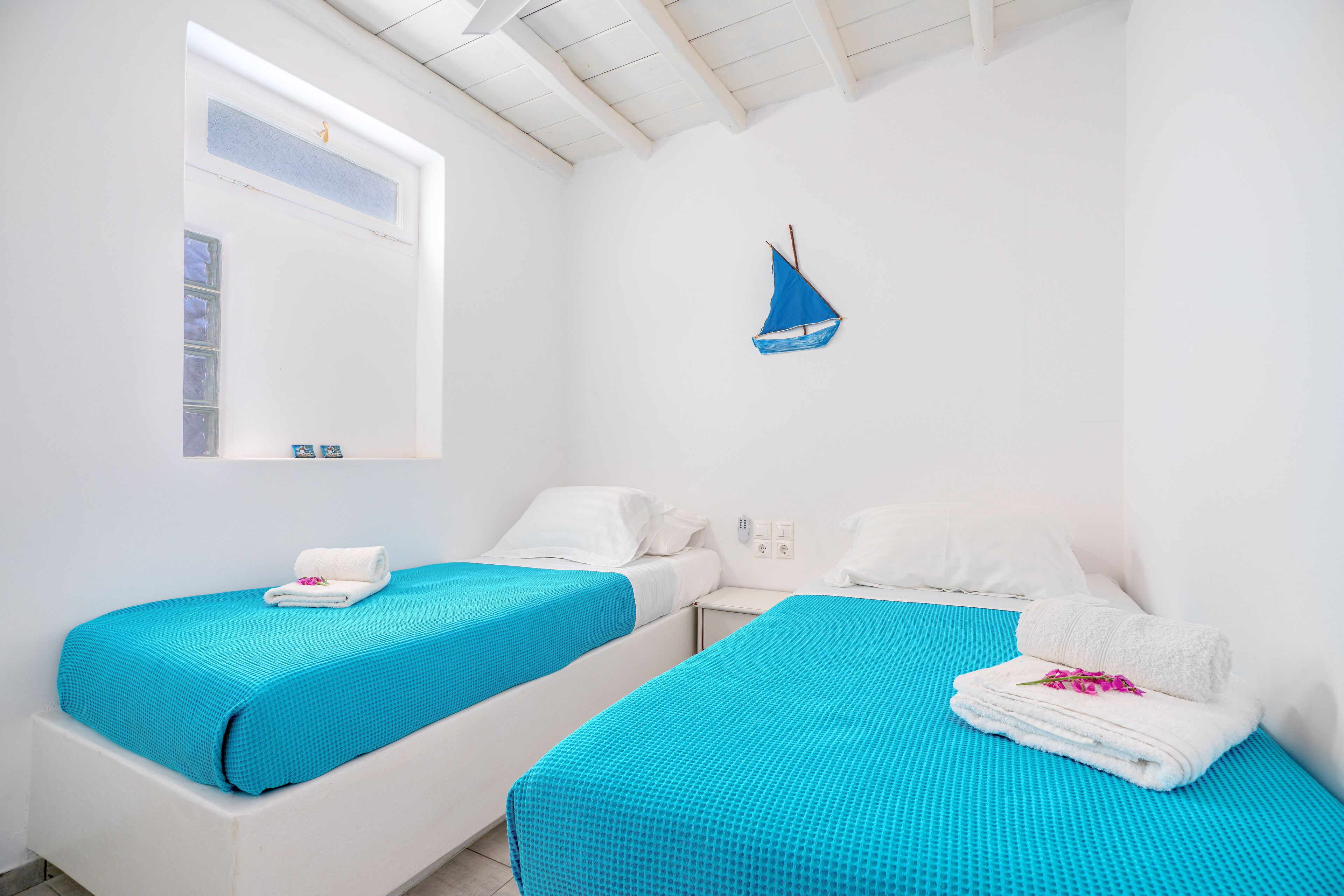 A room with two beds, each with a blue bedspread and white pillows. There is a small sailboat decoration on the wall.