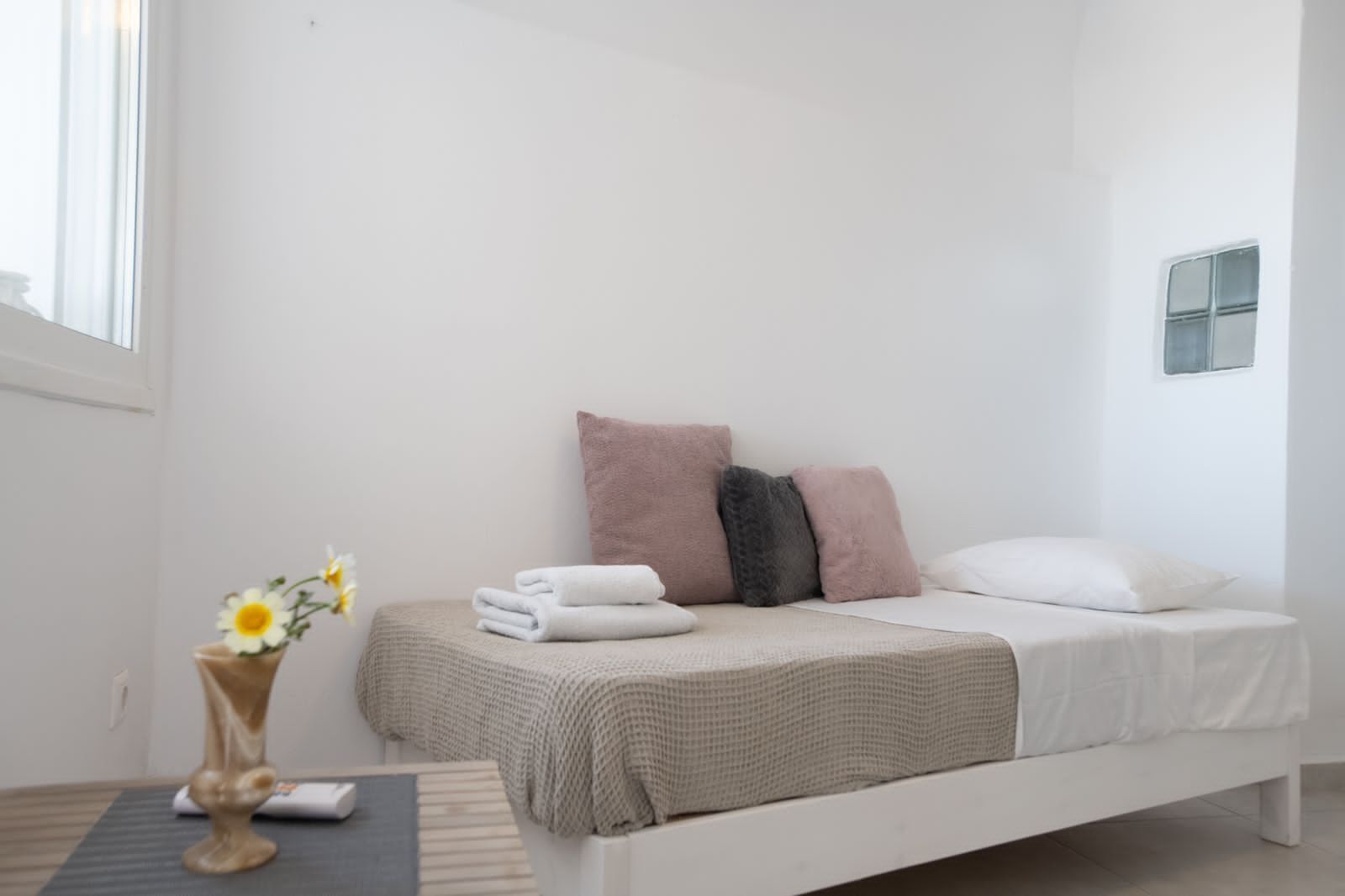 Een minimalistische slaapkamer met een bed, kussens, een nachtkastje met een vaas en een raam.