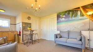A living room with a grey sofa, a dining table with stools, a chandelier, and a beach-themed wall mural.