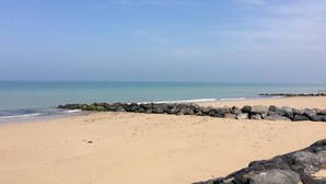 Ein sandiger Strand mit einem steinernen Wellenbrecher im Vordergrund und ruhigem, blauem Wasser im Hintergrund.