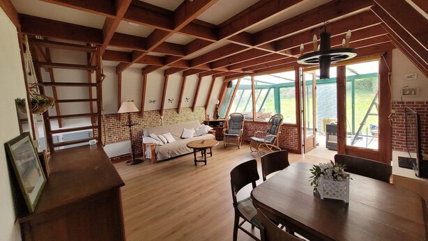 Ein geräumiges Loft mit freiliegenden Holzbalken, einer Couch, zwei Sesseln, einem Esstisch und Blick ins Freie durch große Fenster.