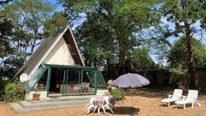 Eine kleine, blockhausartige Hütte mit Glasdoor und Fenstern, umgeben von Bäumen. Zwei weiße Lounge-Stühle und ein violetter Sonnenschirm sind vorhanden.