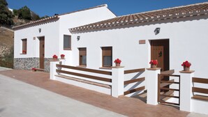 A white building with a tiled roof, wooden doors, and windows with shutters. There are potted plants with red flowers on the porch. - Villa La Noria, 15 Pers, Pool, view of the ocean and the Andalusian mountains (AN)