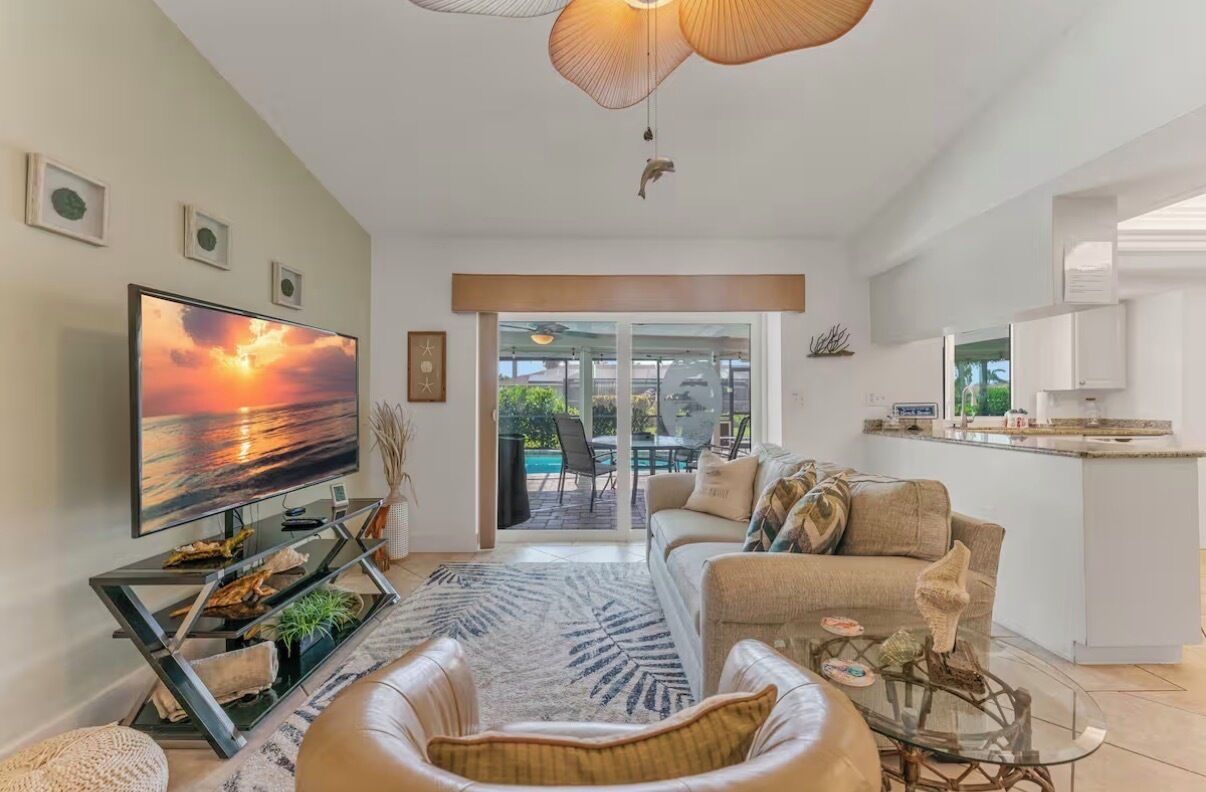 Ein modernes Wohnzimmer mit einem großen Flachbildfernseher, einer Couch, einem Couchtisch und einem Esstisch mit Glasplatte. Ein großes Schiebeglasfenster führt zu einer Außenpatio-Terrasse.