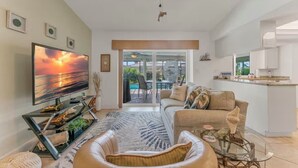 A modern living room with a large flat-screen TV, a sofa, a coffee table, and a glass-top dining table. There is a sliding glass door leading to an outdoor patio area.