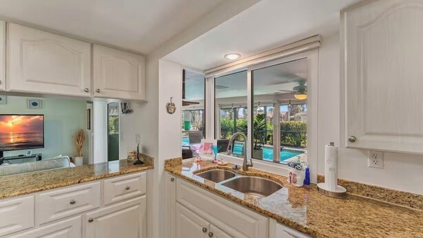 A kitchen with white cabinets, granite countertops, a stainless steel sink, and a window overlooking a swimming pool.