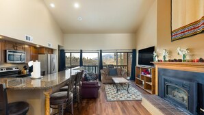 A spacious living area with a kitchen, bar stools, a fireplace, and a large window overlooking a mountainous landscape. - Cozy Top Floor Condo w/ Incredible Mountain Views from Private Balcony! (Silverthorne)