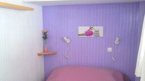 Una habitación con una cama, un taburete de madera, un teléfono de pared y una estantería con una flor.