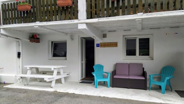 Un edificio blanco con un balcón, un cartel que dice "MARMOTTE", una mesa de picnic blanca, dos sillas turquesas y un sofá morado.