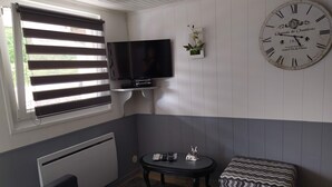 Una habitación con un reloj de pared grande, televisor, una mesita con un adorno y una ventana con persianas.