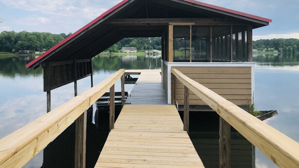 A wooden dock leading to a covered pavilion with a red roof, overlooking a calm lake with a house in the background.