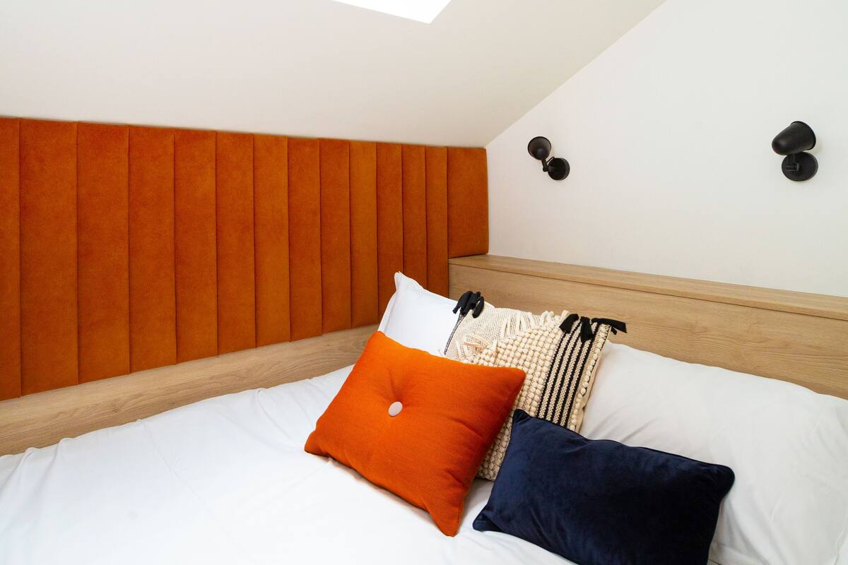 A bedroom with a sloped ceiling, a skylight, a bed with white bedding, orange and dark blue pillows, and a wooden headboard.