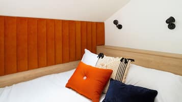 A bedroom with a sloped ceiling, a skylight, a bed with white bedding, orange and dark blue pillows, and a wooden headboard.