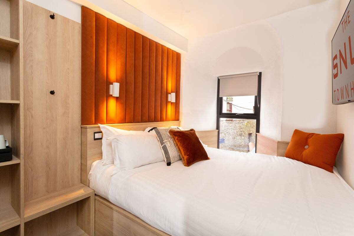 A modern hotel room with a wooden headboard, white bedding, and a view of the outside through a window.