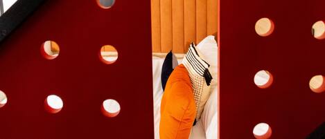 Un interior moderno con una pared de acento rojo, una cama con ropa de cama blanca y un cojín naranja, y un diseño de listones de madera.