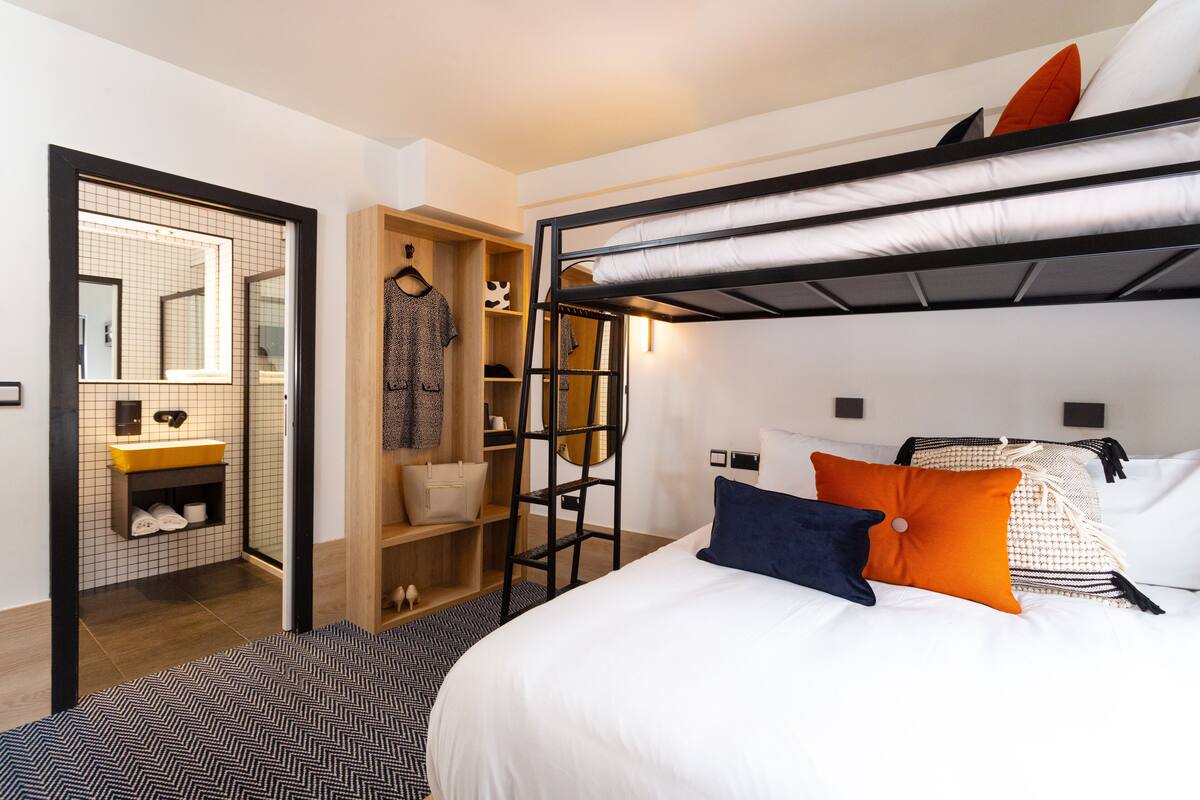 A bunk bed with a ladder, a wardrobe, a mirror, and a bathroom with a sink and towel rack.