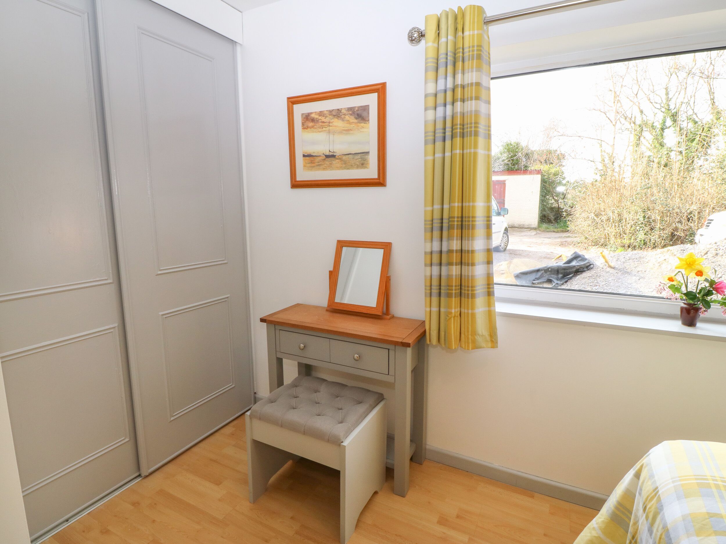A bedroom with a wooden desk, a small stool, a framed picture, and a window with curtains.