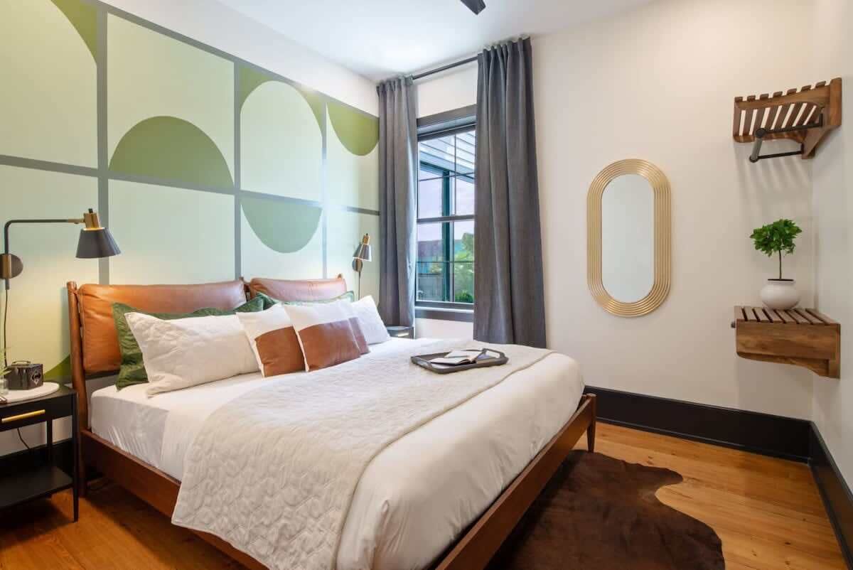 A modern bedroom with a large bed, a nightstand, a lamp, a window with curtains, and a wall-mounted shelf with a plant.