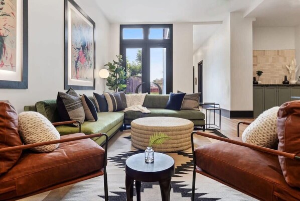 Een moderne woonkamer met een hoekbank, twee leren fauteuils, een rond hocker en een salontafel. Er is een groot raam met uitzicht op de omgeving, kunstwerken aan de wanden en een keuken op de achtergrond.