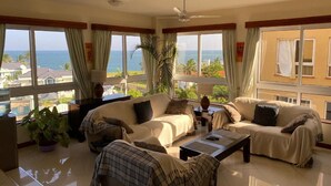 A spacious living room with two sofas, a coffee table, and a ceiling fan. Large windows offer a view of the ocean and surrounding buildings. - Stay.Plus APA Apartments Nyali (Mombasa)