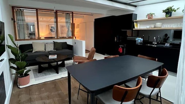 A compact living space with a dining area, a sofa set, a kitchen, and a view through a large window. - Design Led 1 bed in Cosmopolitan Queens Park (London)