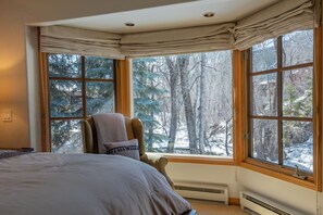 Relax in this serene room with a large bed and forest view, featuring a window with a valance and a cozy chair.