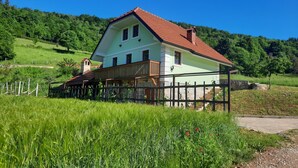 Una casa a due piani con tetto in tegole rosse, immersa in campi verdi rigogliosi e un cielo azzurro limpido.