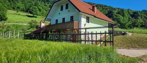A two-story house with a red-tiled roof, surrounded by lush green fields and a clear blue sky.