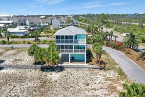 A two-story beachfront house with a balcony, surrounded by palm trees and a sandy beach.