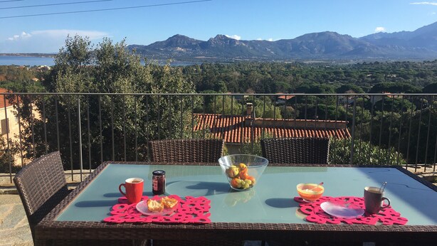 A table with a clear view of mountains, a tablecloth, and a bowl of fruit.
