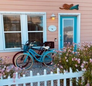 Un vélo turquoise est garé devant une maison avec une clôture blanche et des fleurs roses.