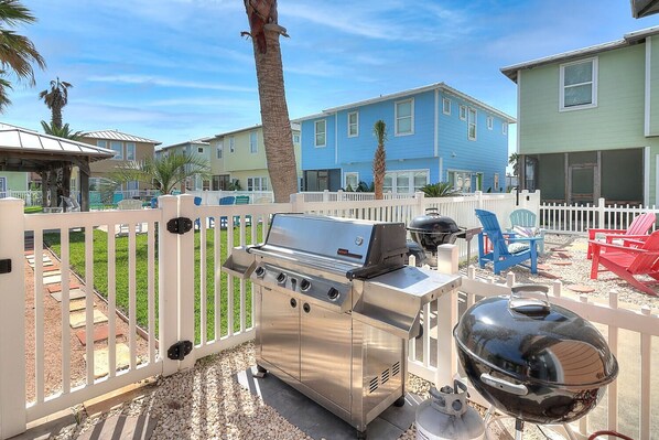 A backyard with a stainless steel grill, a barbecue grill, and a gas tank. There are white fences, a palm tree, and a blue house in the background. - Gorgeous, pet friendly, coastal cottage within walking distance to the restauran (Port Aransas)