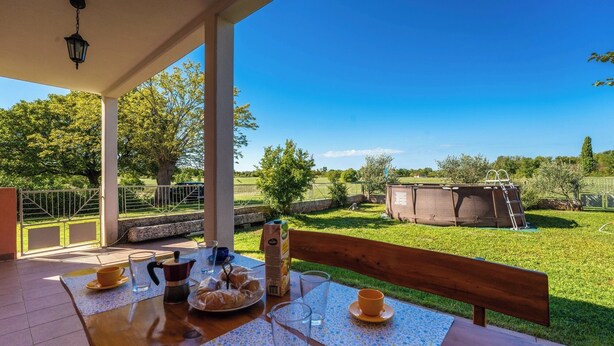 An outdoor dining area with a table set for two, a bench, and a view of a grassy field and a swimming pool. - Ines in Barban  - Haus für 4-5 Personen (Barban)