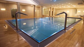 Una piscina interior moderna con paredes de vidrio transparente, suelo de madera y una fuente central.