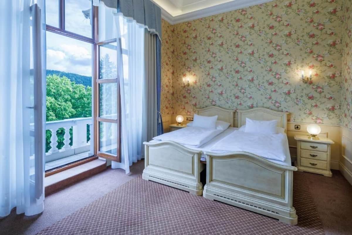 A bedroom with a large window, a double bed, two bedside tables, and floral wallpaper.