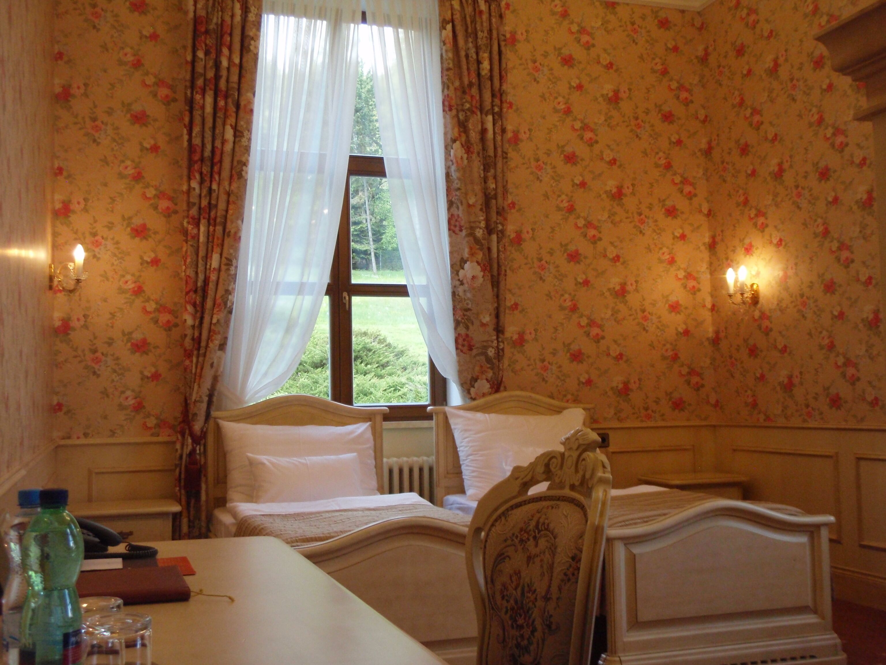 A bedroom with floral wallpaper, a bed with white linens, a wooden headboard, a nightstand with a lamp, and a window with sheer curtains.