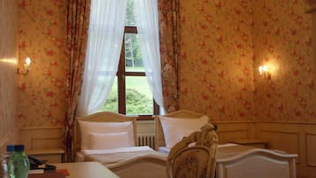 A bedroom with floral wallpaper, a bed with white linens, a wooden headboard, a nightstand with a lamp, and a window with sheer curtains.
