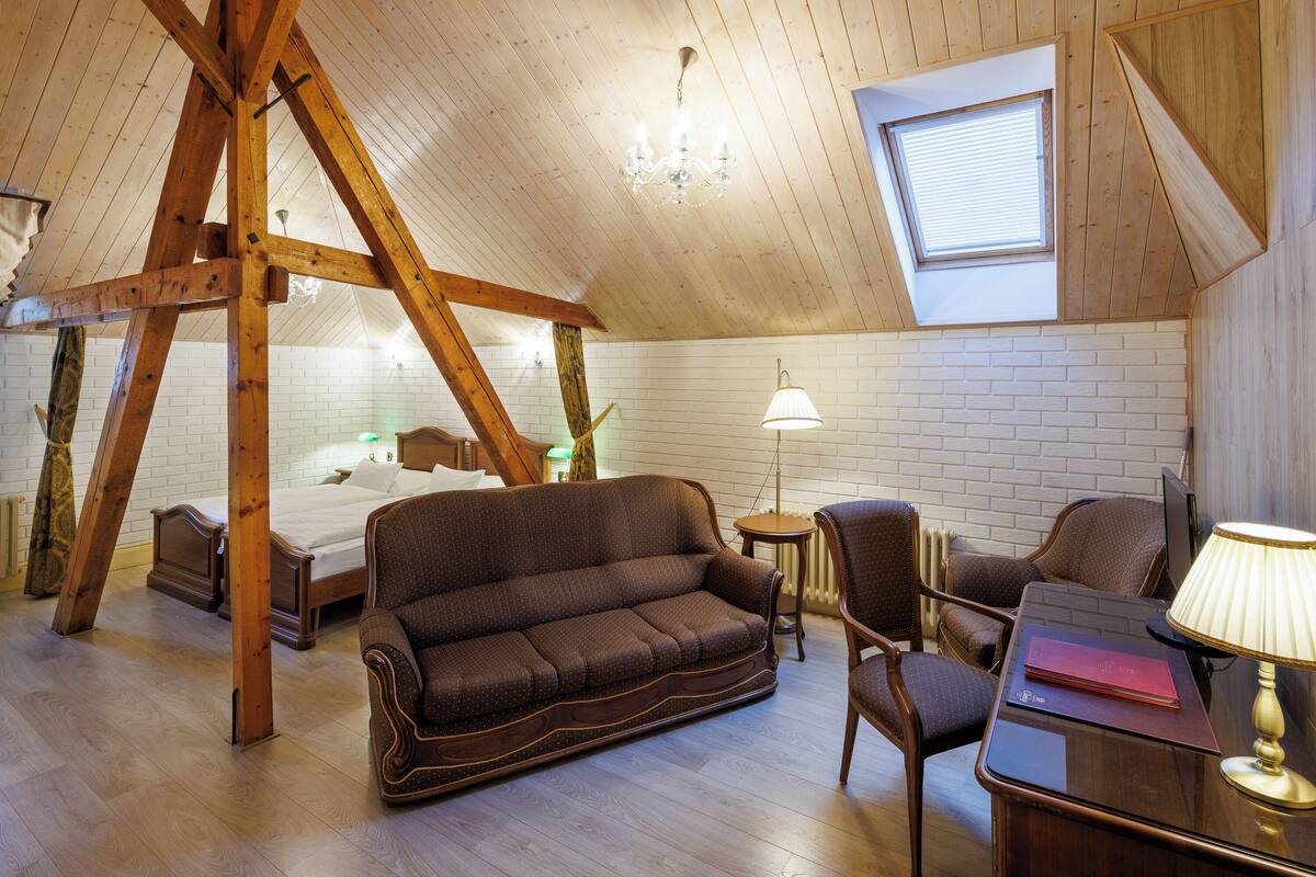 A cozy attic bedroom with a wooden ceiling, a sloped roof, a bed, a brown sofa, a desk with a lamp, and a window.