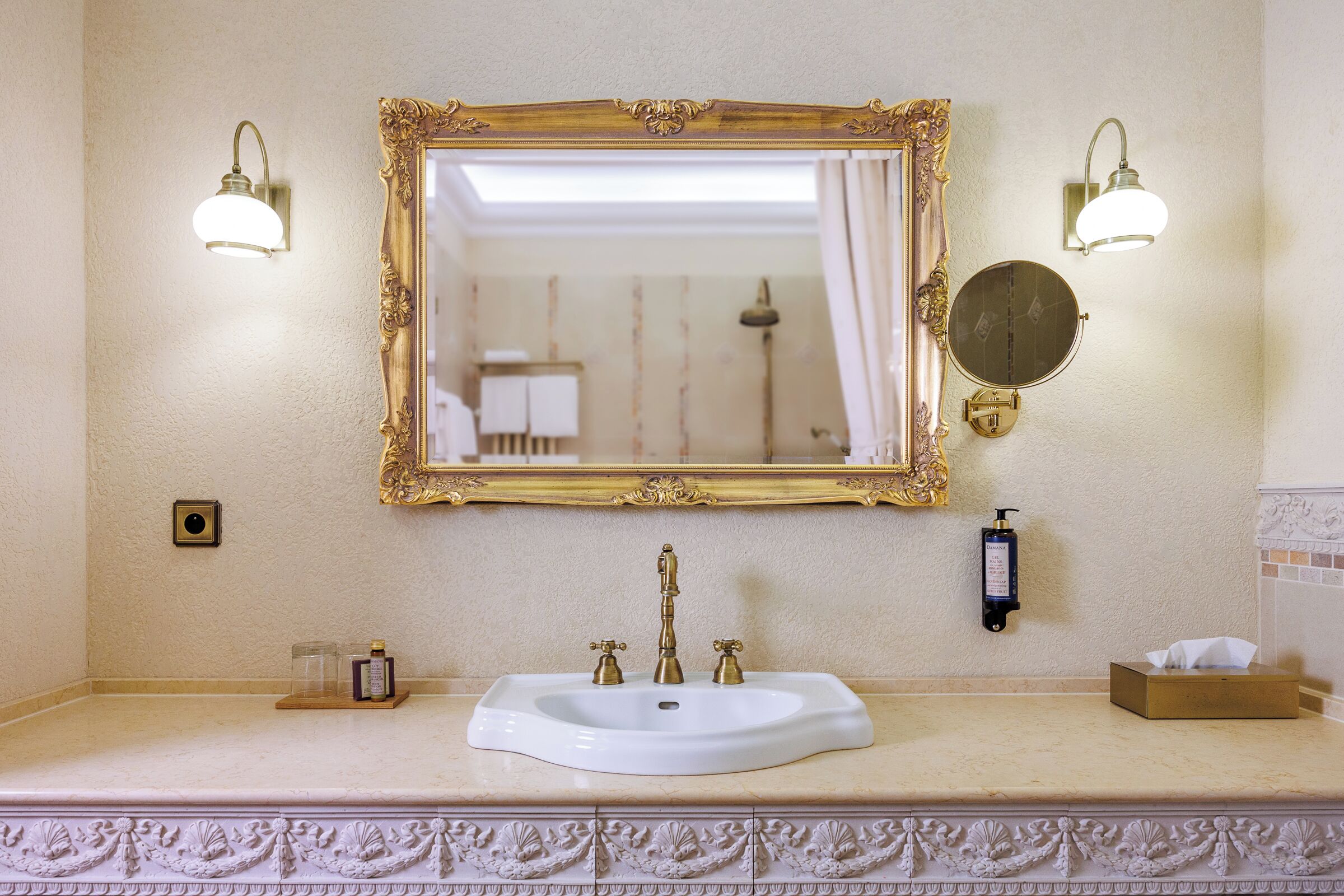 A bathroom with a white sink, gold faucets, and a large ornate mirror. There are two wall-mounted light fixtures and a round mirror below the main mirror.