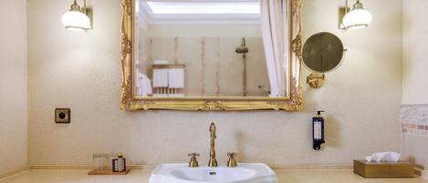 A bathroom with a white sink, gold faucets, and a large ornate mirror. There are two wall-mounted light fixtures and a round mirror below the main mirror.