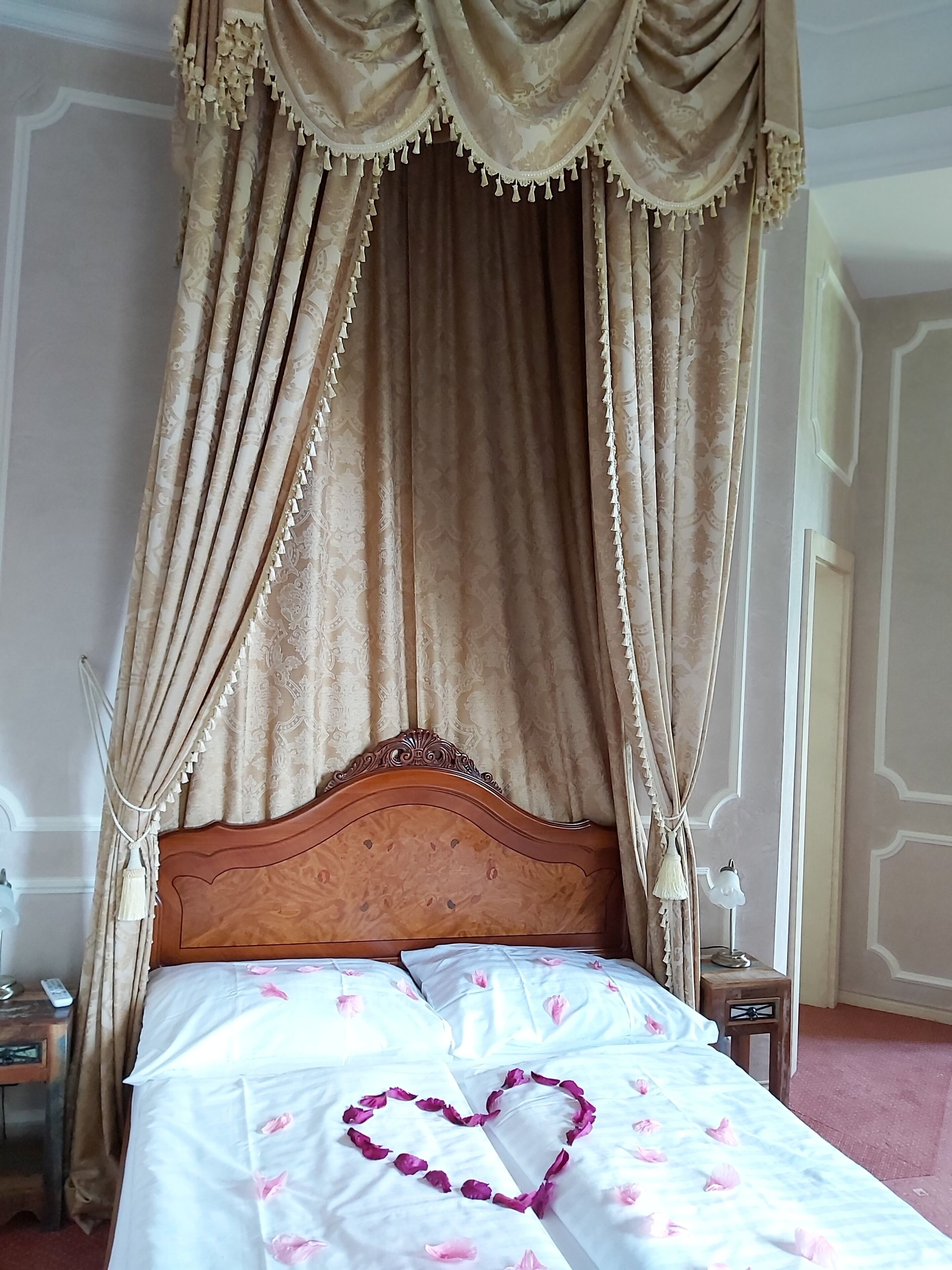 A bed with a heart-shaped decoration made of rose petals, a wooden headboard, and a canopy with tassels.